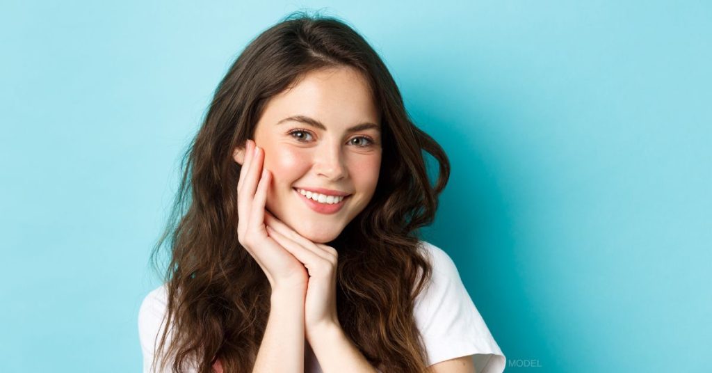 Woman standing against blue backdrop smiling with hand on face (MODEL)